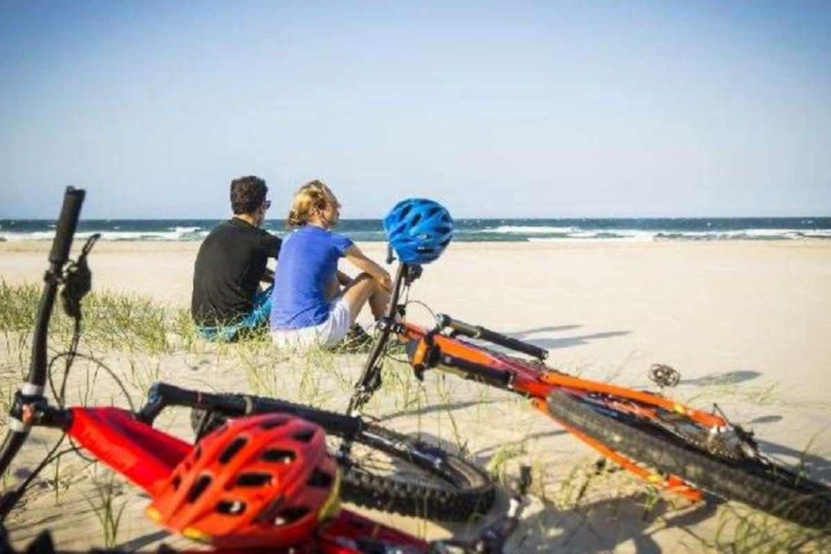 Two riders sitting on the sand at a Noosa surf beach with their bikes beside them