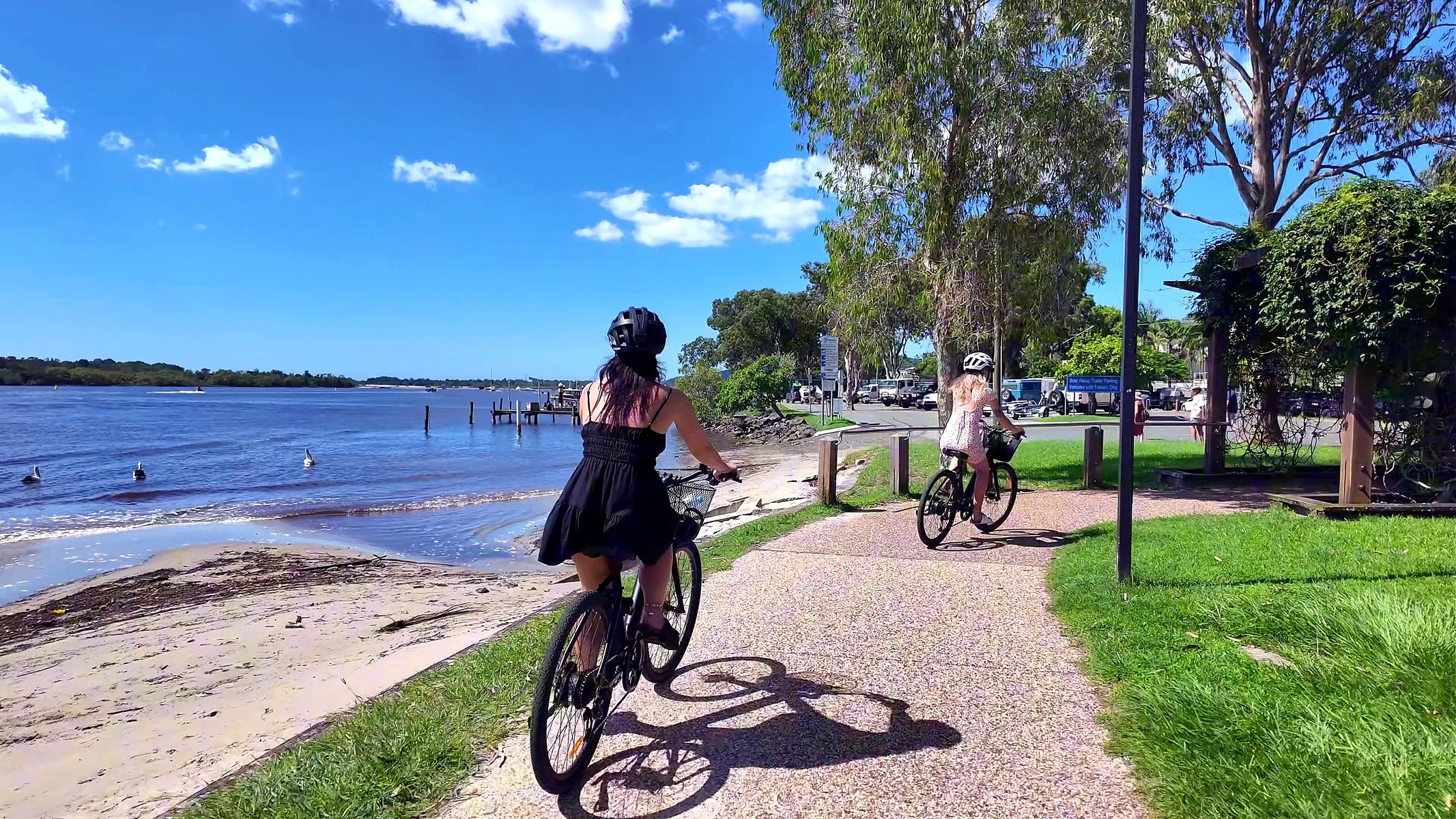 Two riders on e-bikes on the Noosa River bike path, pelicans on the water beside them