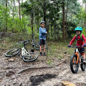 Young rider on an orange bike riding a forest trail as a friend watches on